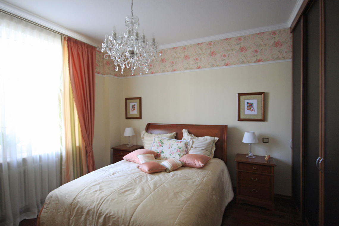 Стены спальни оформлены обоями цвета шампань с широким бордюром с цветочным рисунком под потолком. Потолок украшает прозрачная хрустальная люстра.
